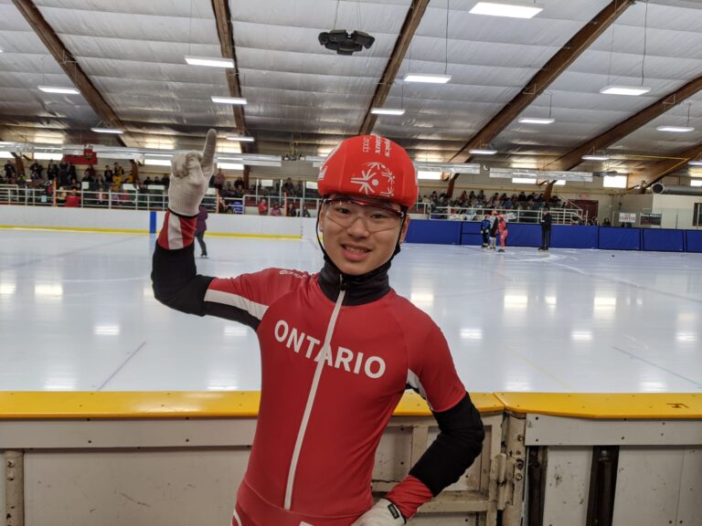 Speed skater Ezekiel Liu holding up his index finger beside an indoor skating rink